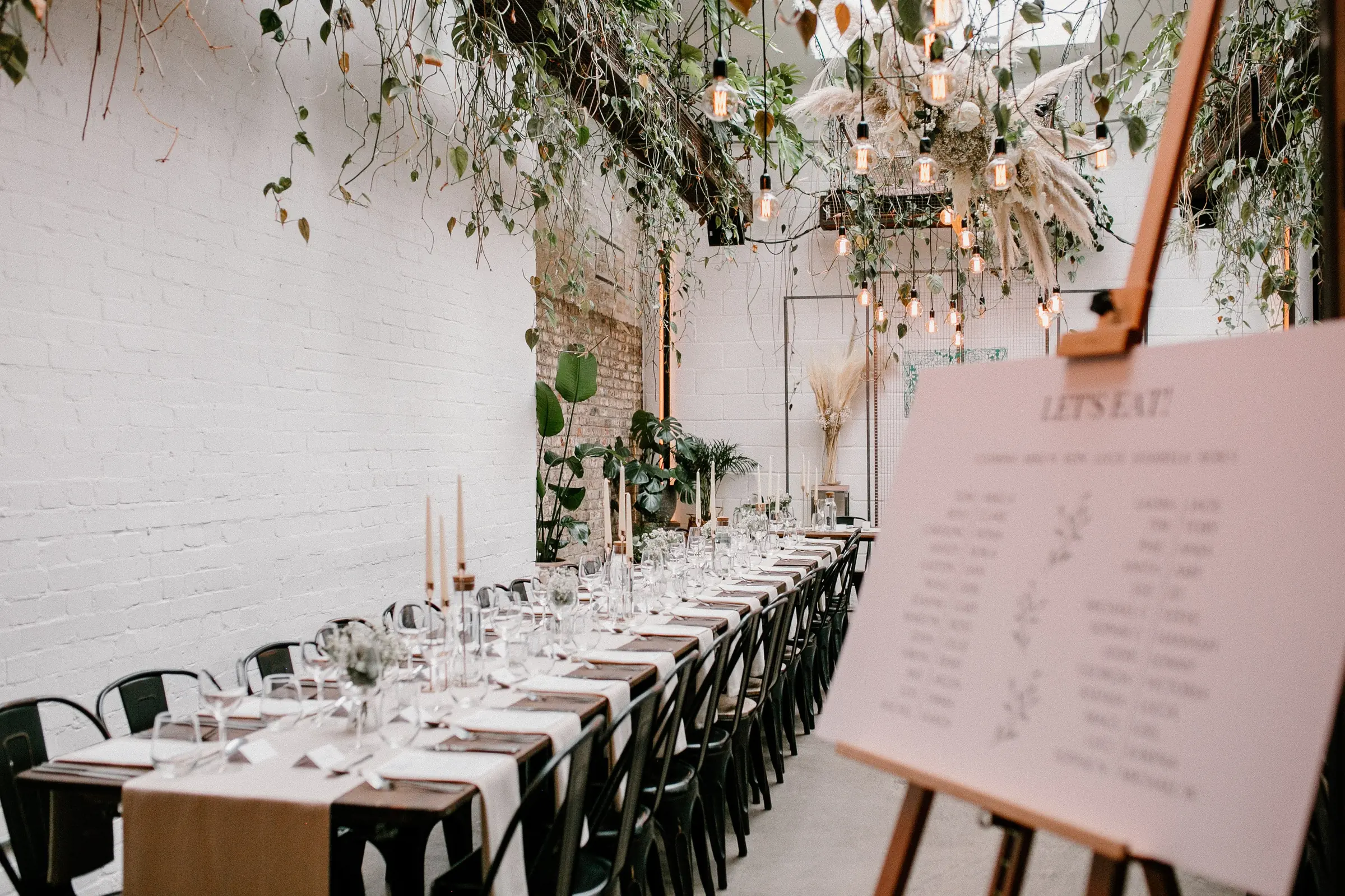 Long candlelit banquet table at The Hackney set for dinner, with hanging greenery and Edison bulb chandelier